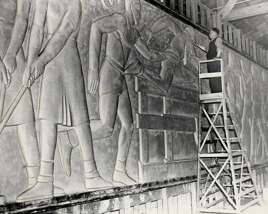 A black-and-white photo of a man standing on a tall wooden ladder and working on an elaborate wall sculpture.