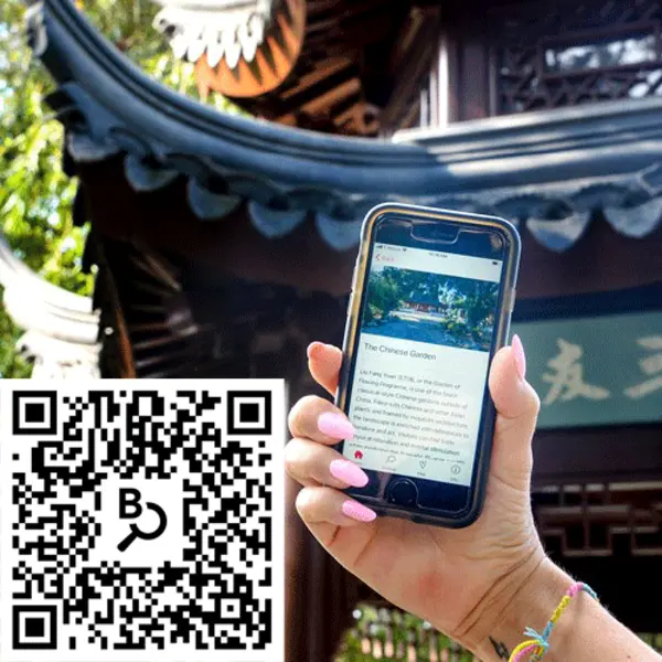 Hand holding up an iPhone in front of a pavilion in the Chinese Garden with a QR code on the bottom left. 