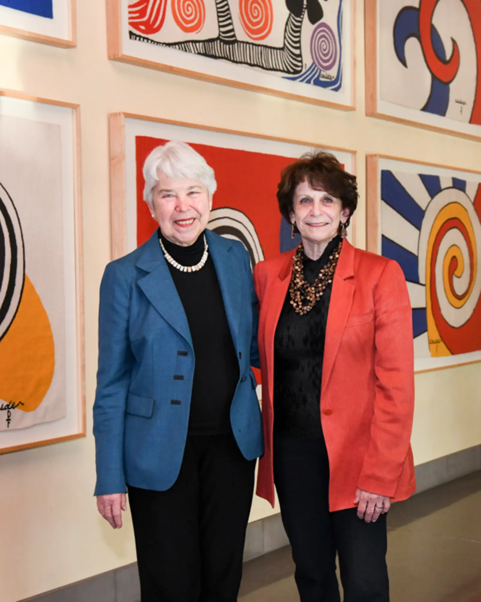 Two people stand together in front of a wall of art in primary colors.