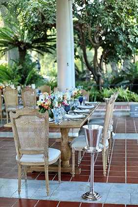 Wooden outdoor dining tables and whicker-backed dining chairs.	