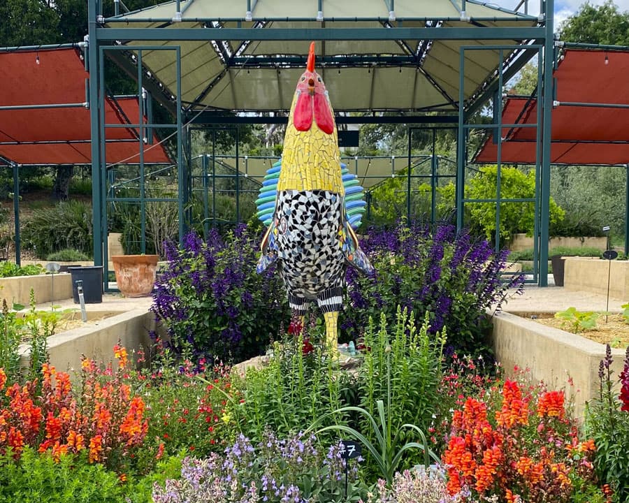 A large sculpture of a rooster, covered in mosaic tiles, surrounded by flowers.