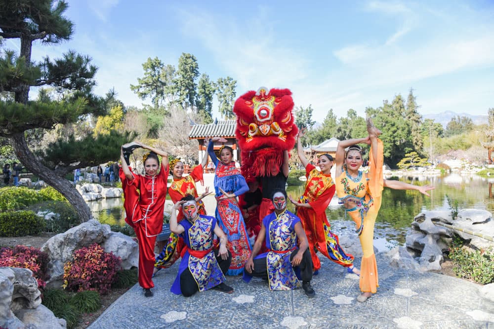 A group of performers in colorful outfits pose outdoors in a garden.