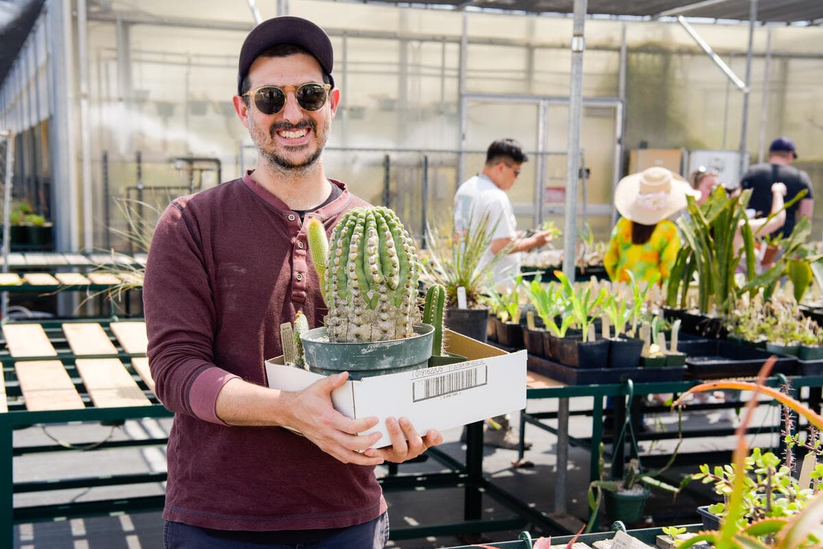 A smiling person in sunglasses holds a box with potted plants.