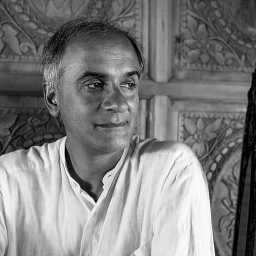 A black-and-white photo of a person in a white shirt, seated in front of a wood carved wall.