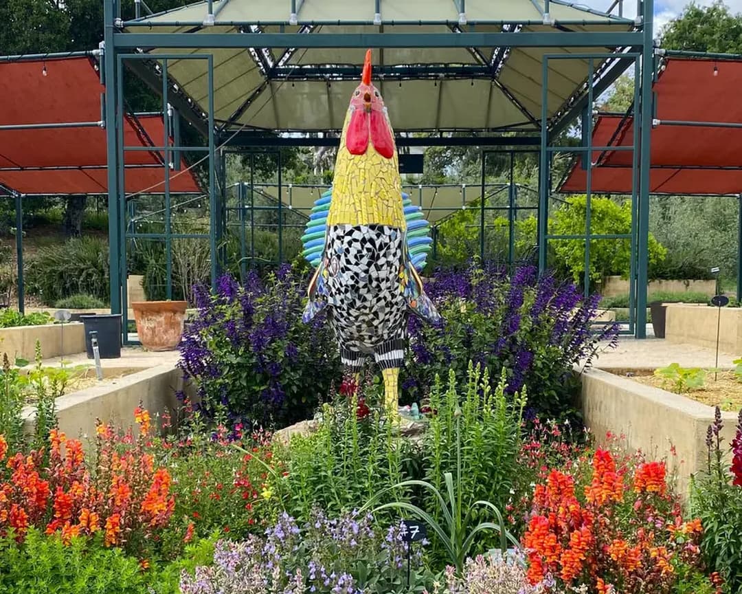 A large rooster sculpture made of colorful tiles in a garden of flowering plants.