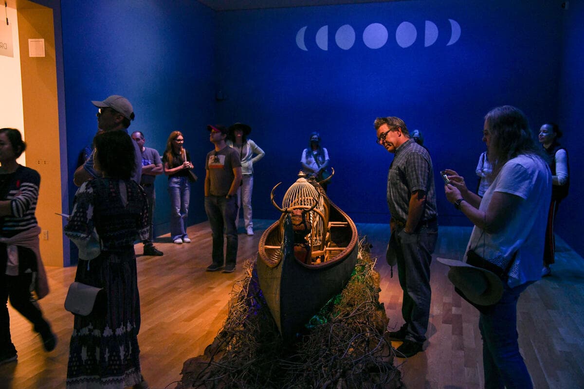 People stand in a gallery with blue walls, looking at a sculpture made from found objects, including a canoe, chairs, and cages.