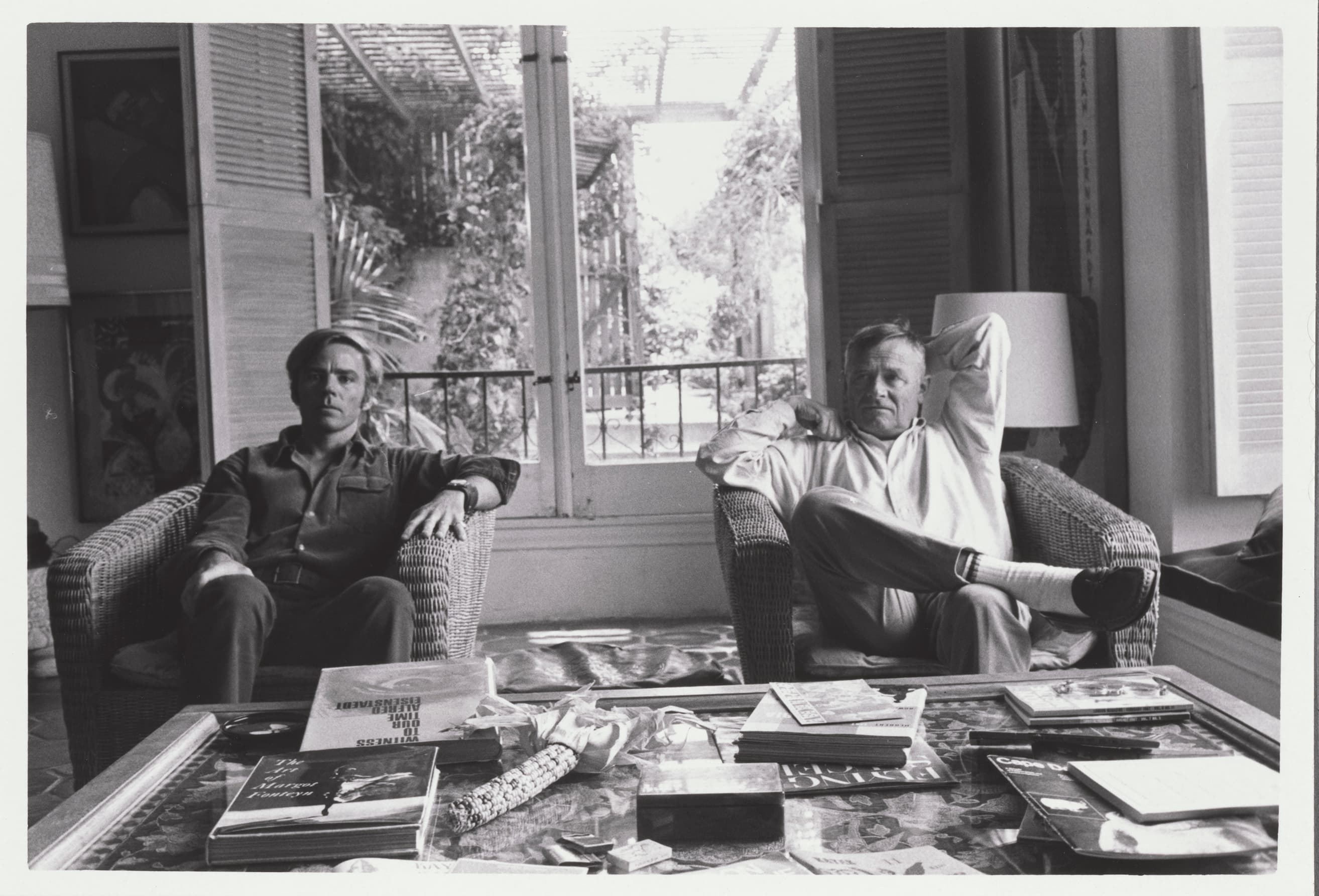 A black-and-white photo of two men in a living room, sitting behind a coffee table filled with books.