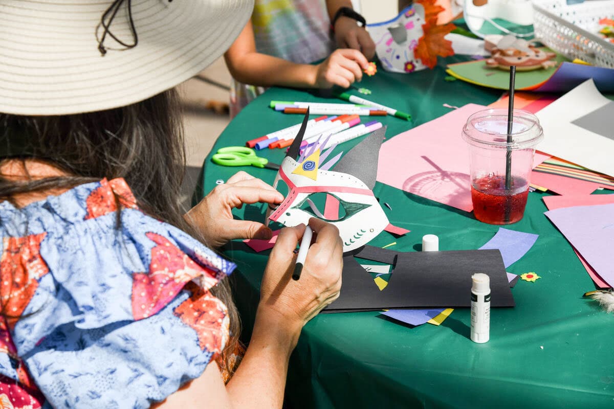 Over-the-shoulder view of a person decorating a paper mask on a green table filled with art supplies.
