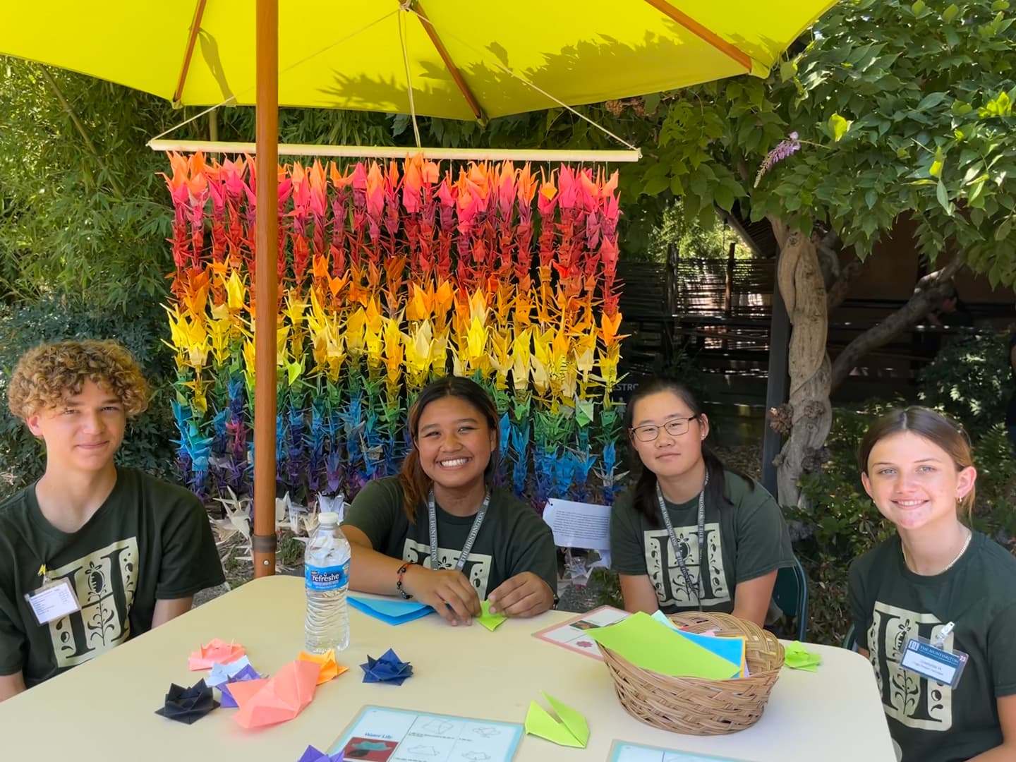 Four young people sit at a table with craft supplies, in front of a curtain of folded paper cranes.