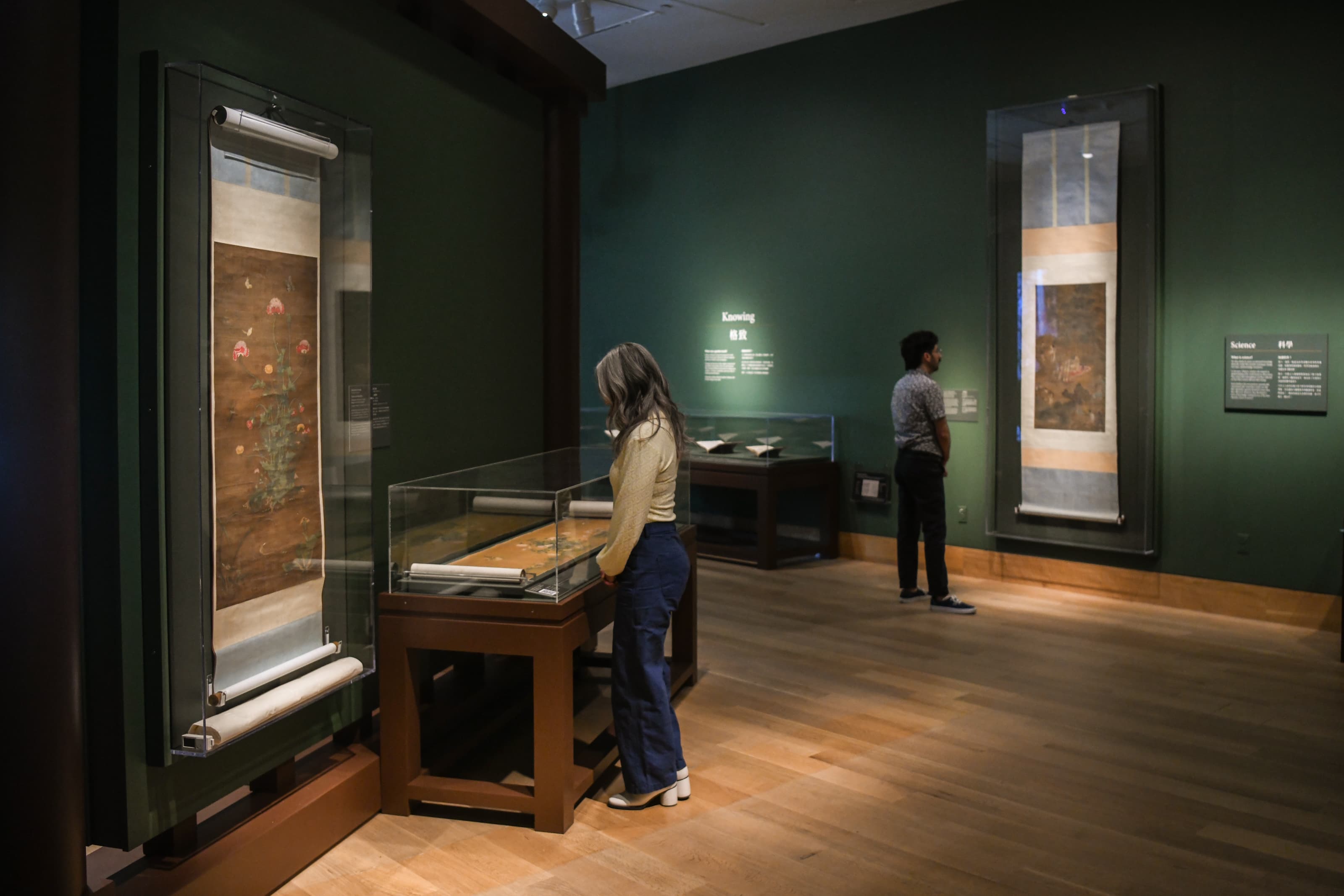 Two people stand in an art gallery with green walls, with large traditional Chinese scroll paintings on display.