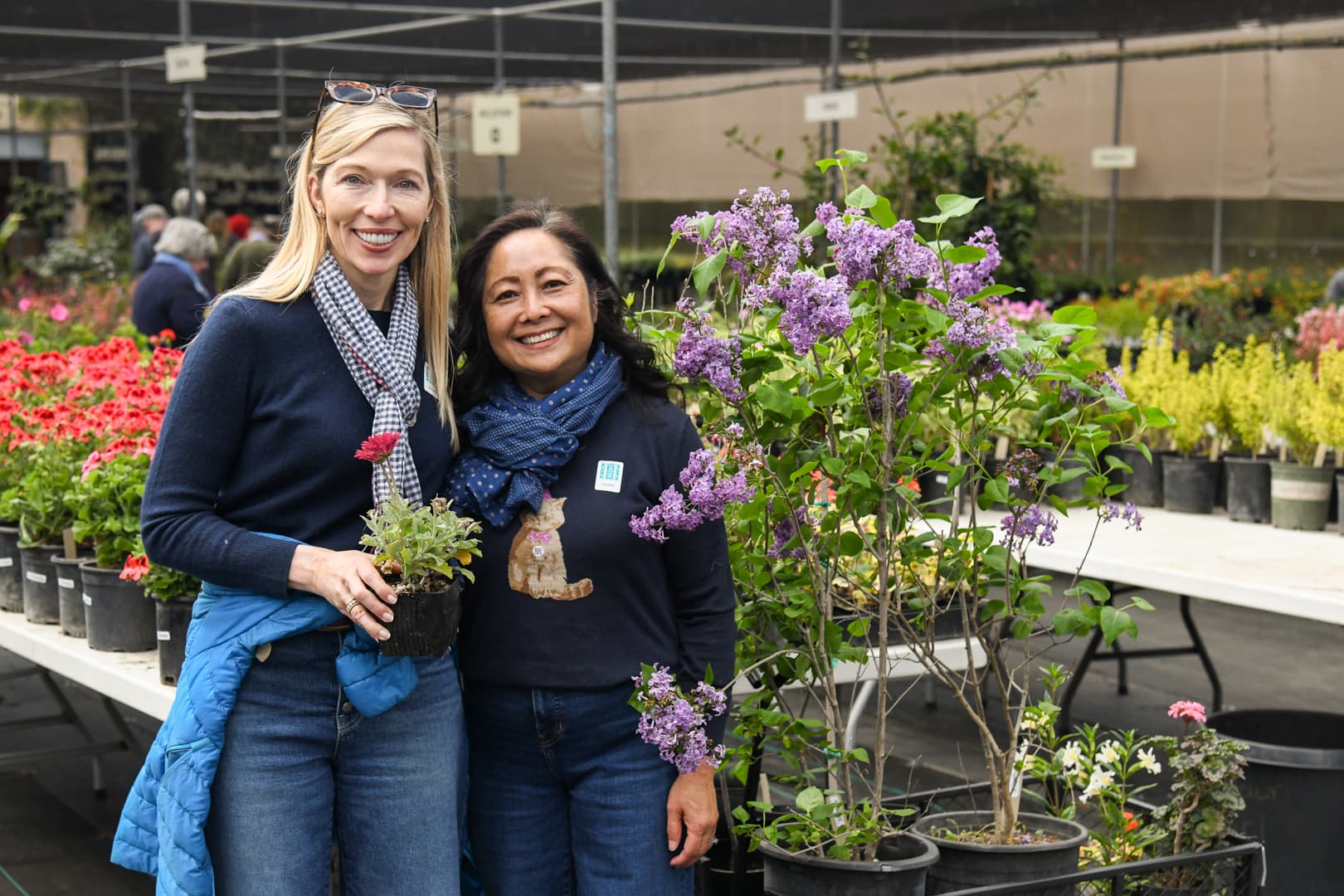 Two smiling people stand near each other, in front of tables filled with potted plants.
