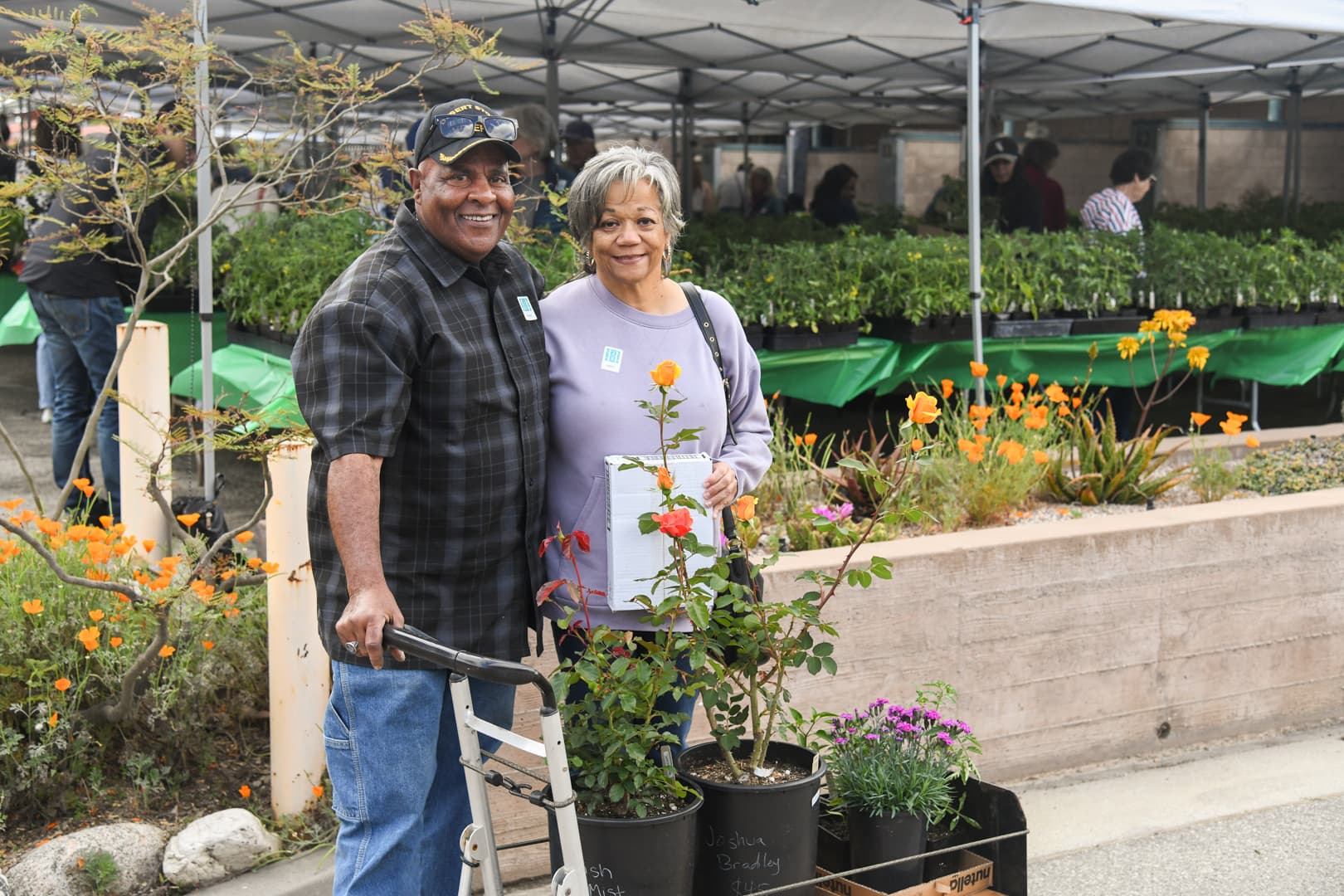 Two smiling people stand near a cart filled with potted plants.