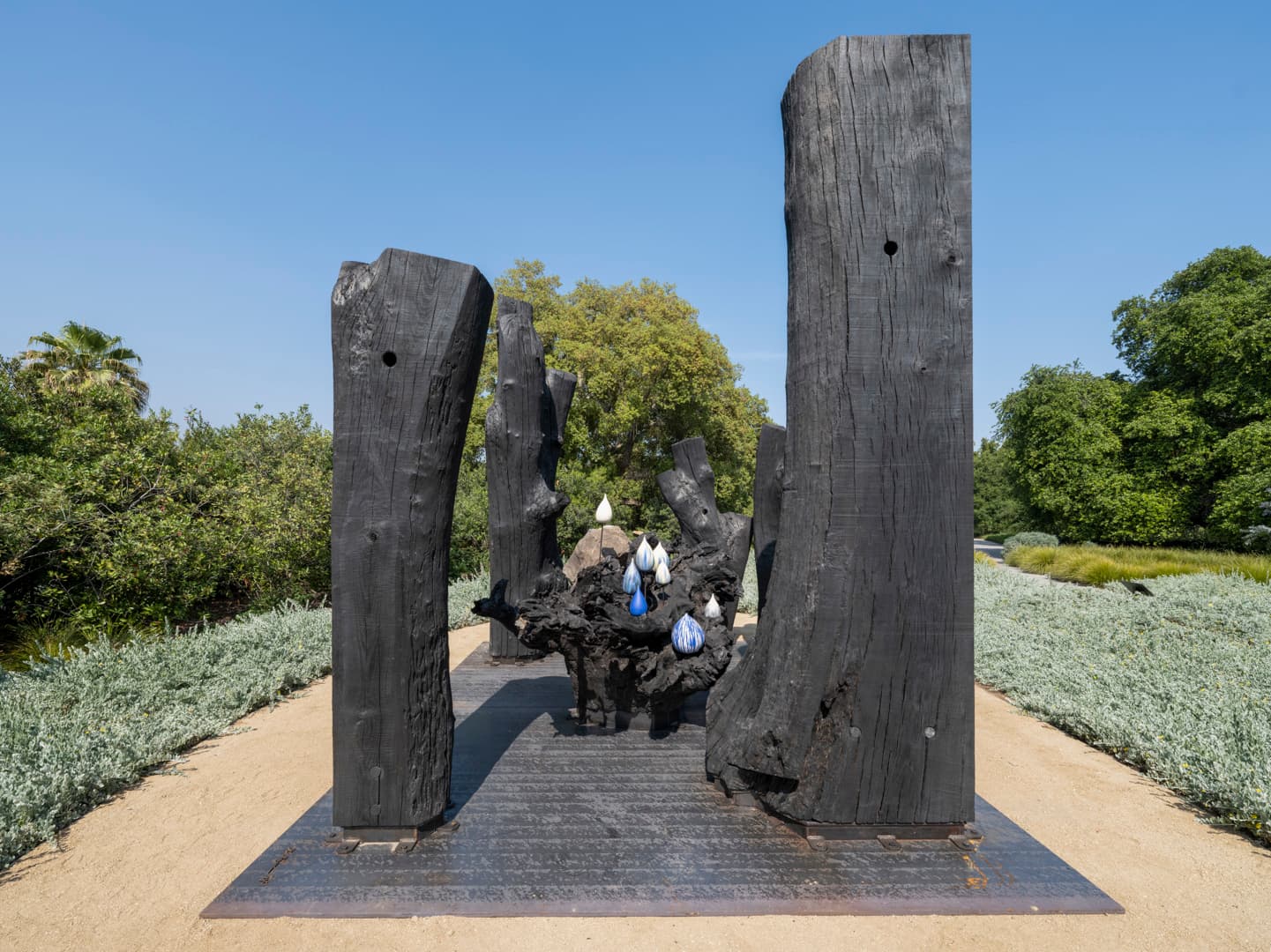 A wood sculpture in a garden, with ceramic droplets at the center.