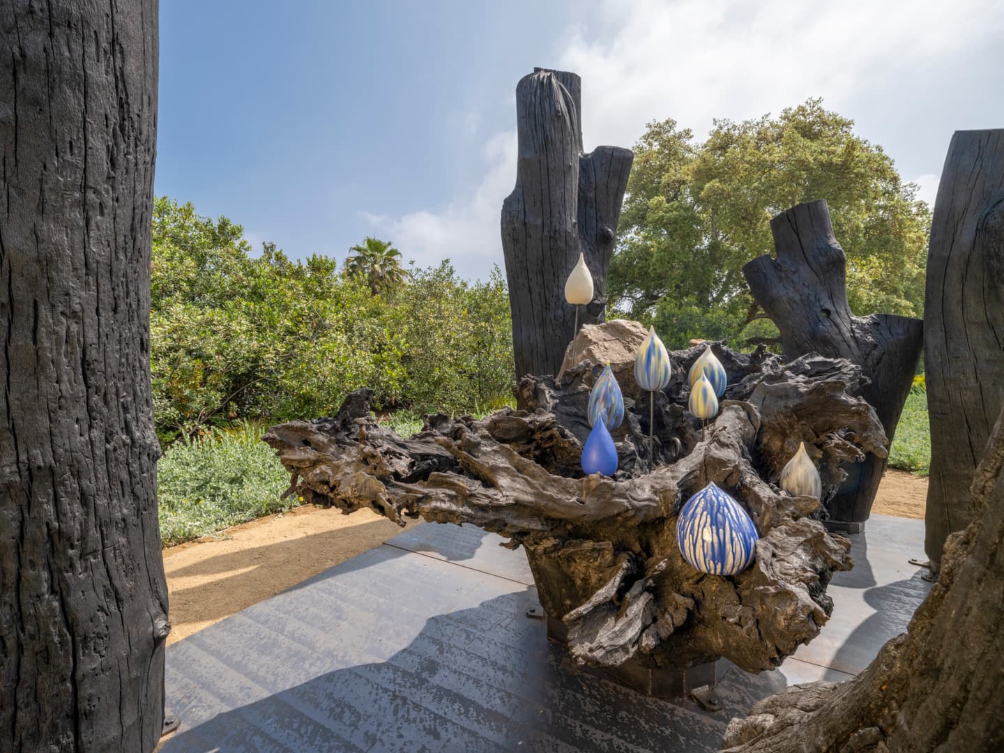 A wood sculpture in a garden, with ceramic droplets at the center.