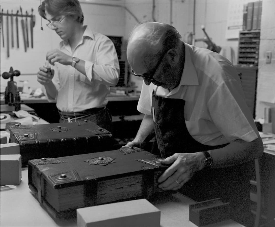 A black and white photo of two people in a workshop at a table with a large book.