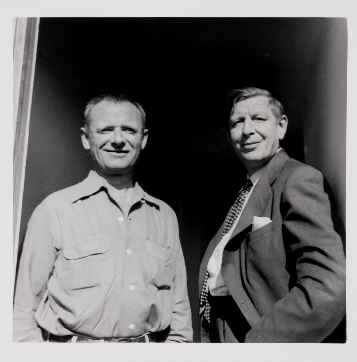 Black-and-white photo of two people looking at the camera, one in a button down shirt, the other in a suit and tie.