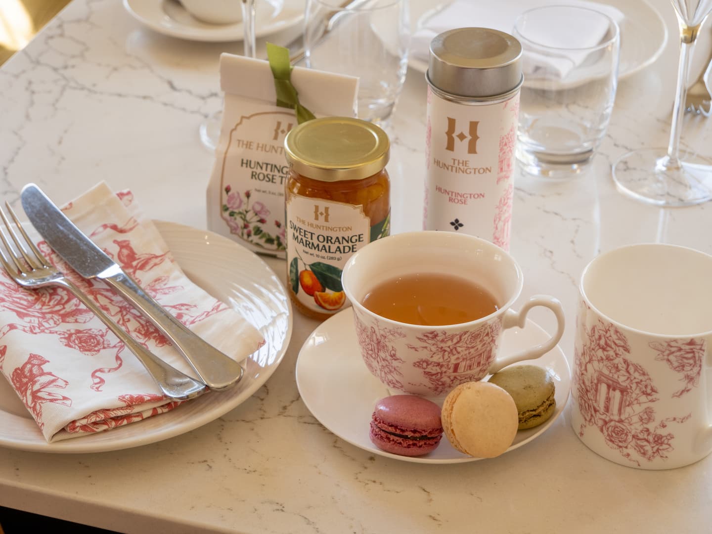 A table setting with serveware in a pink toile pattern, a jar of orange marmalade, and two containers of tea.
