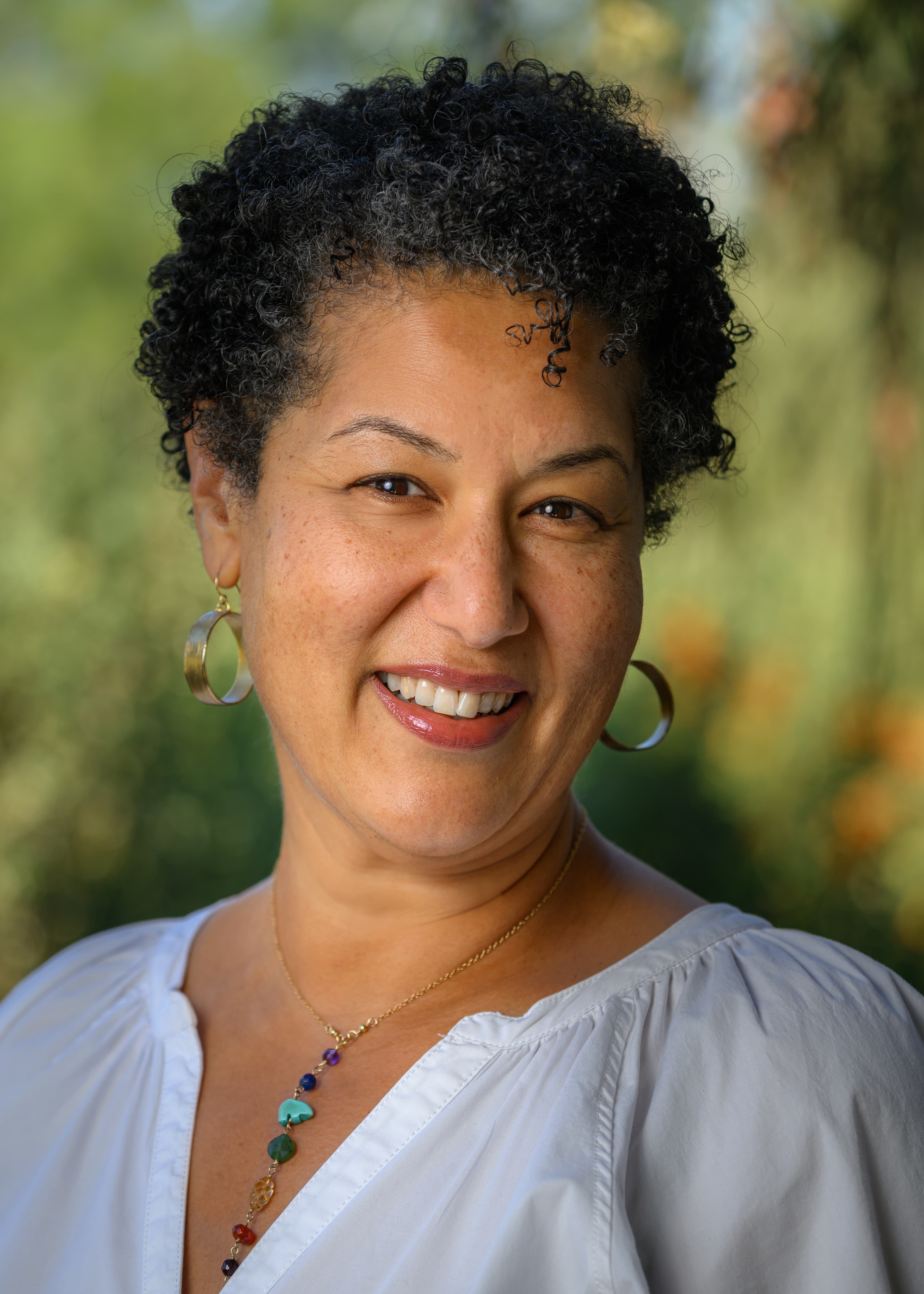 Portrait photo of a person in a white top, crystal necklace and loop earrings, taken outside.