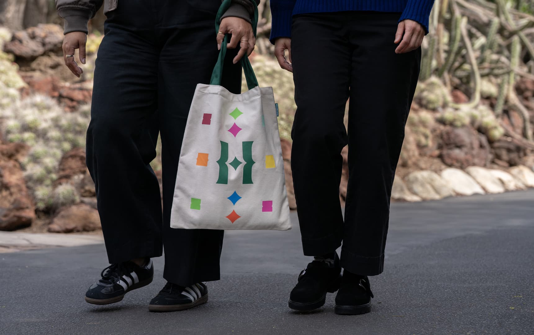 Two people’s legs are visible as they walk on a path in a desert-style garden; one person holds a white tote bag with a colorful design.