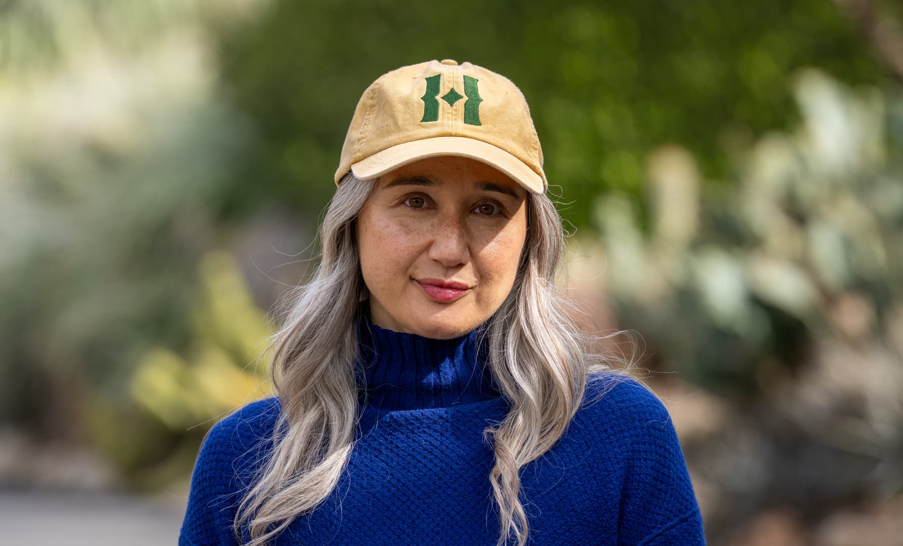 A person with long gray hair wears a blue sweater and a yellow hat with a green logo.