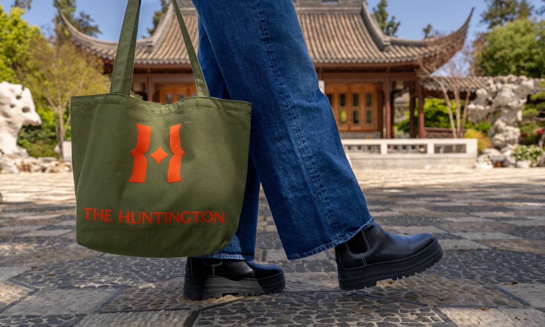 A person holding a green tote bag with orange logos stands in a Chinese-style garden.