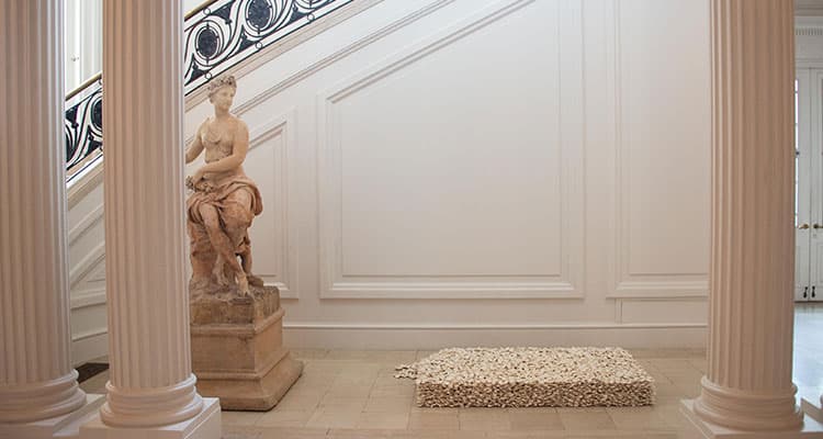 A view of a gallery with a sculpture of a person on a pedestal on the left, on the right a block of ceramic flowers.