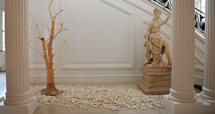 A gallery with a sculpture of a tree on left, a sculpture of a person on a pedestal, the floor in-between is filled with ceramic flowers.