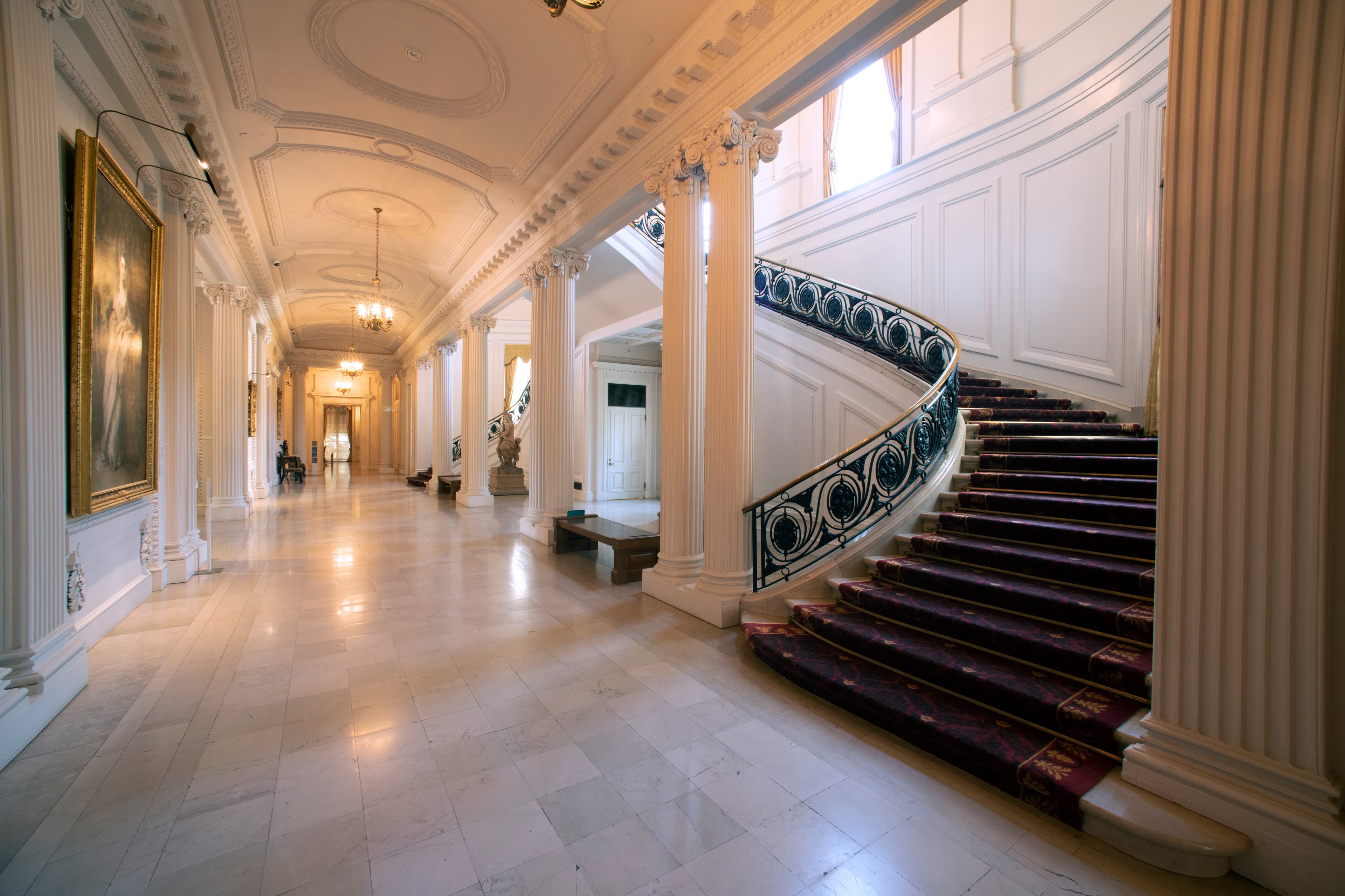 A long hallway in a warmly lit guilded age hallway; A grand staircase leads upwards to the right.