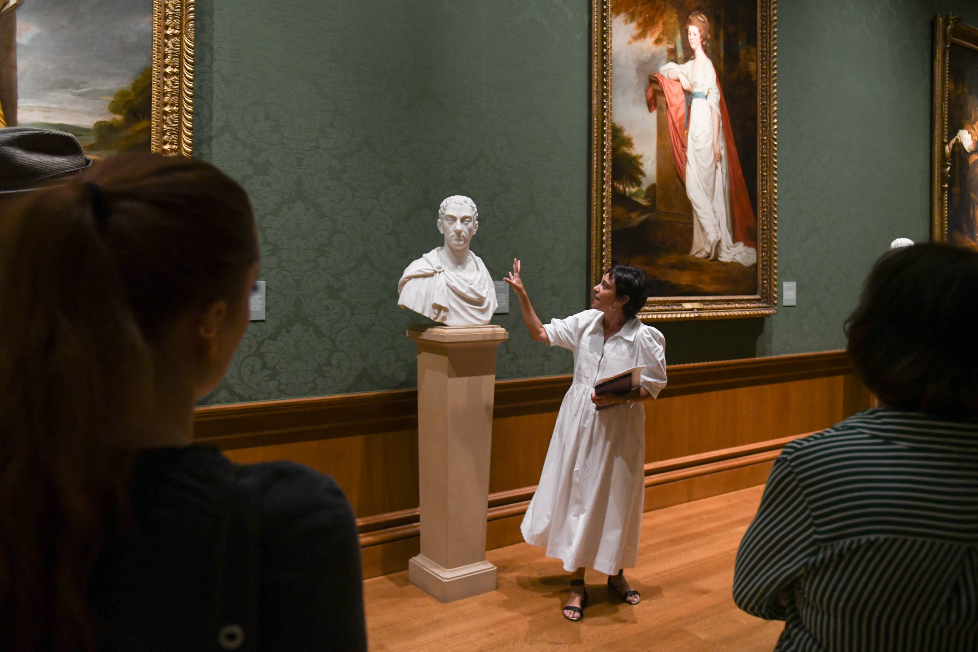 A person stands in front of a group of people discussing a marble statue on a stand in a gallery.