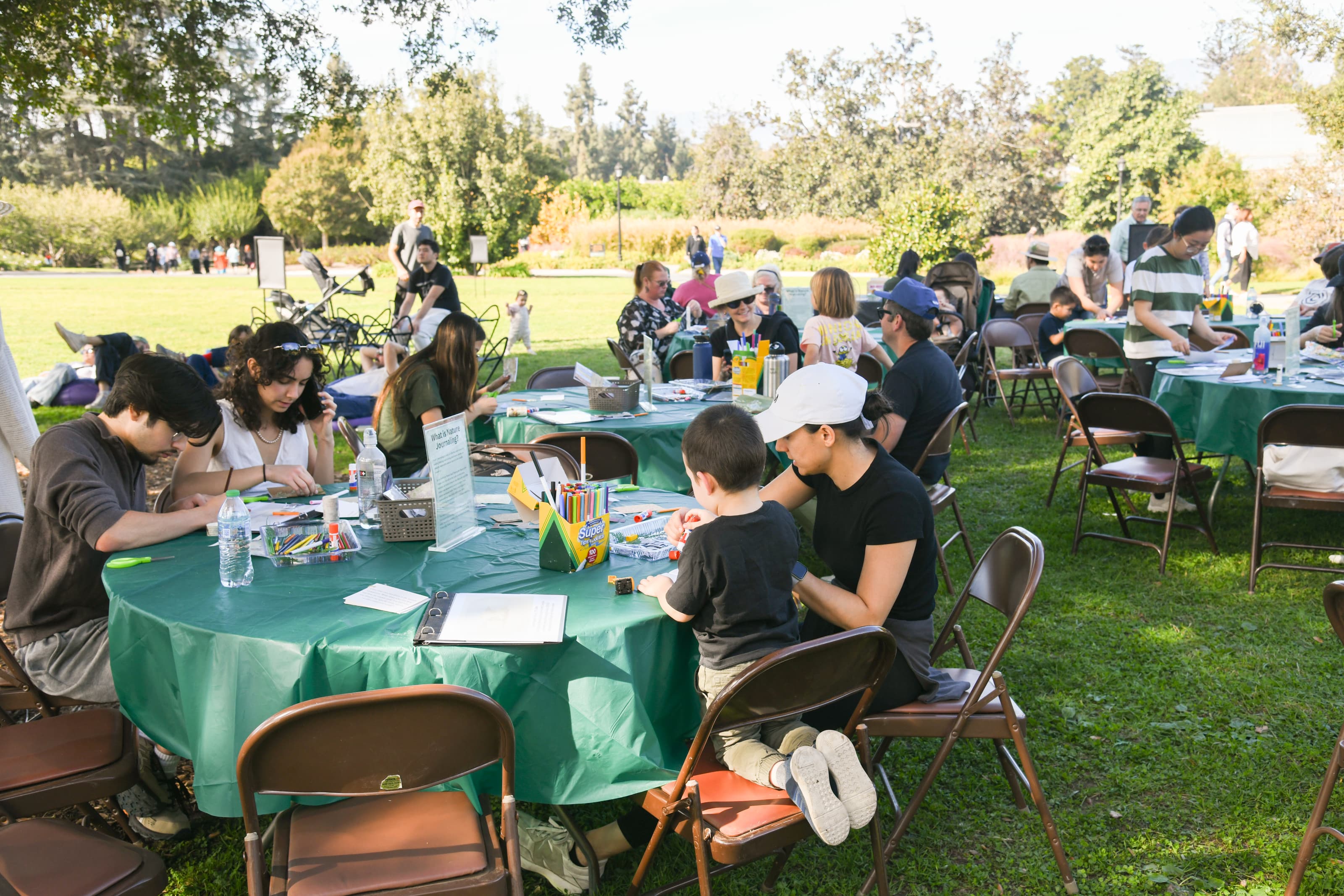 Adults and children create art projects on various outdoor tables in a grassy field.