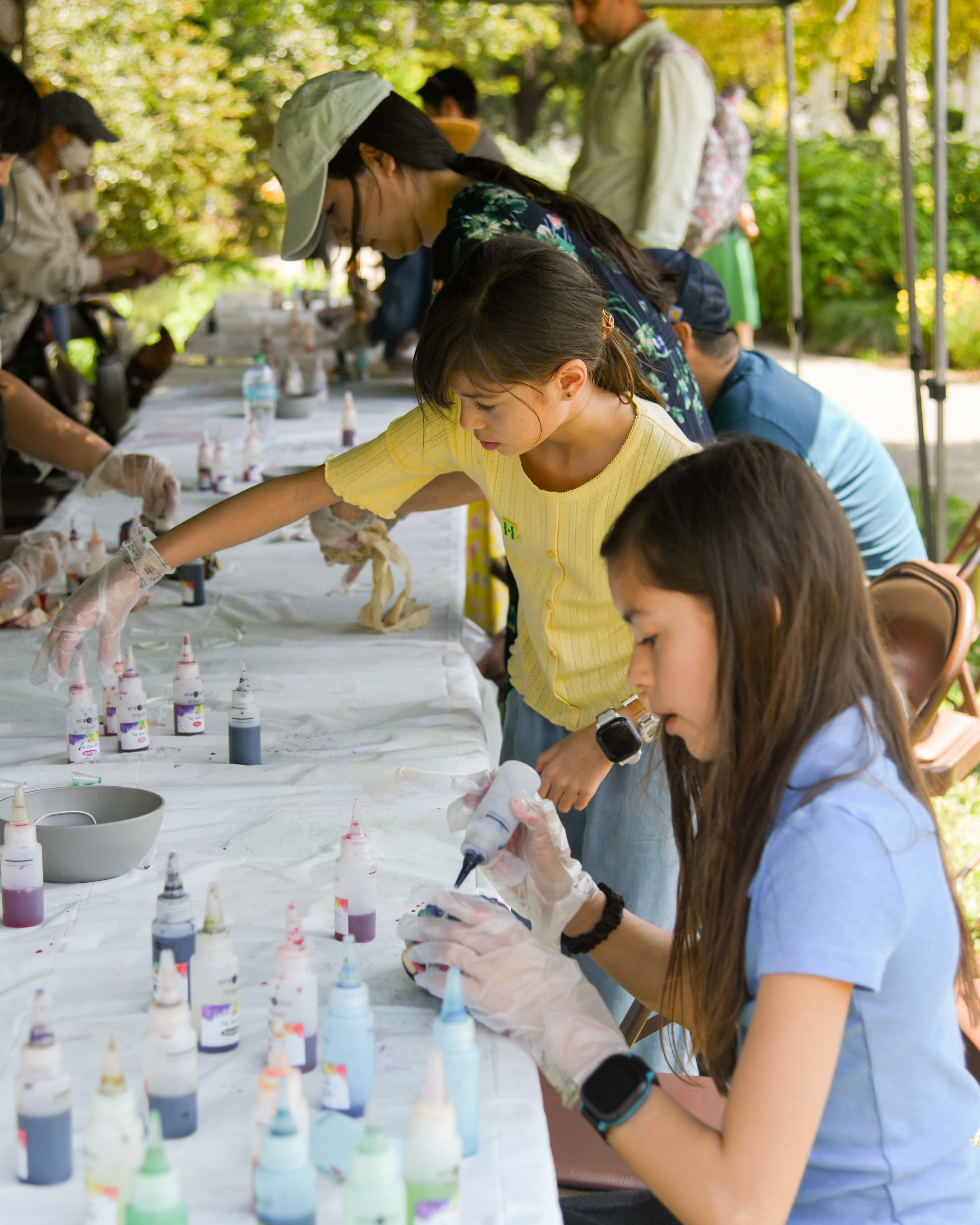 Kids and adults at a long table pick up bottles of different tie-dye colors from a table.