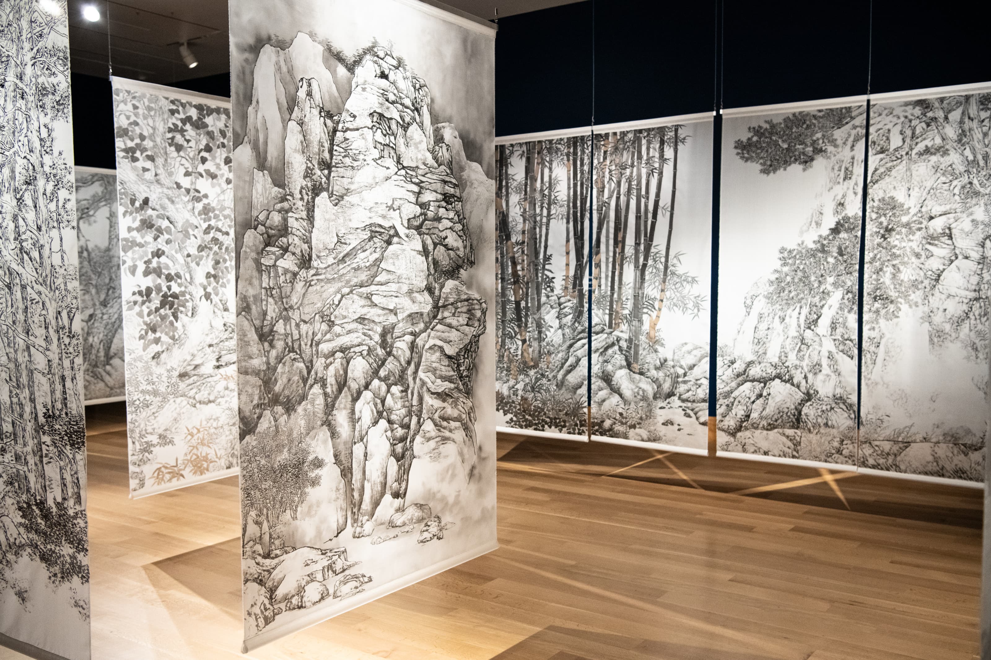 A gallery space with wood floors and dark walls, large silk tapestry paintings hang from the ceiling.
