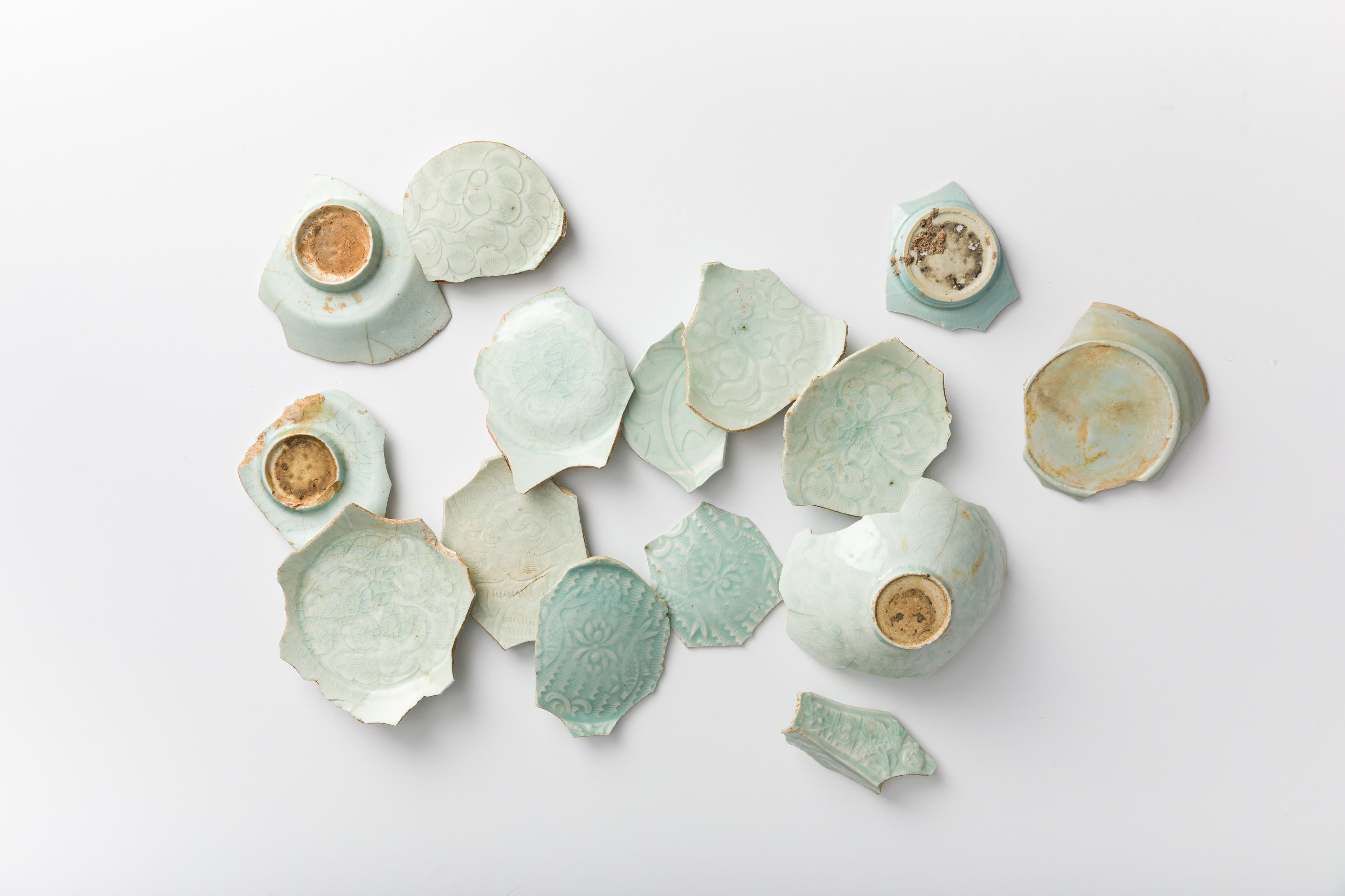 Assorted fragments of pale blue-green glazed pottery, including bowl bases and decorated shards with floral and geometric patterns, displayed on a white background.