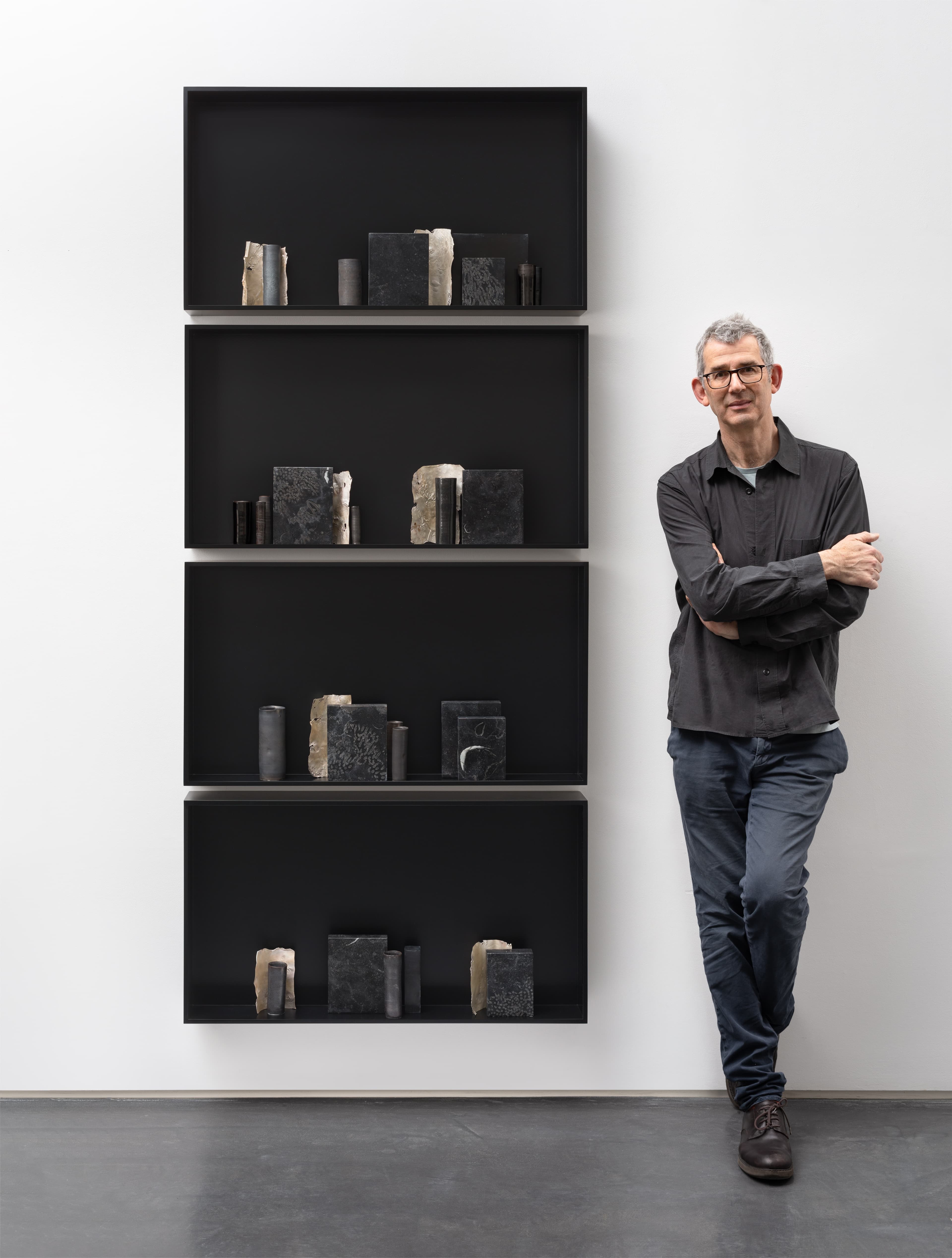 Man with gray hair and glasses standing with arms crossed beside a black, wall-mounted shelving unit displaying book-like objects.