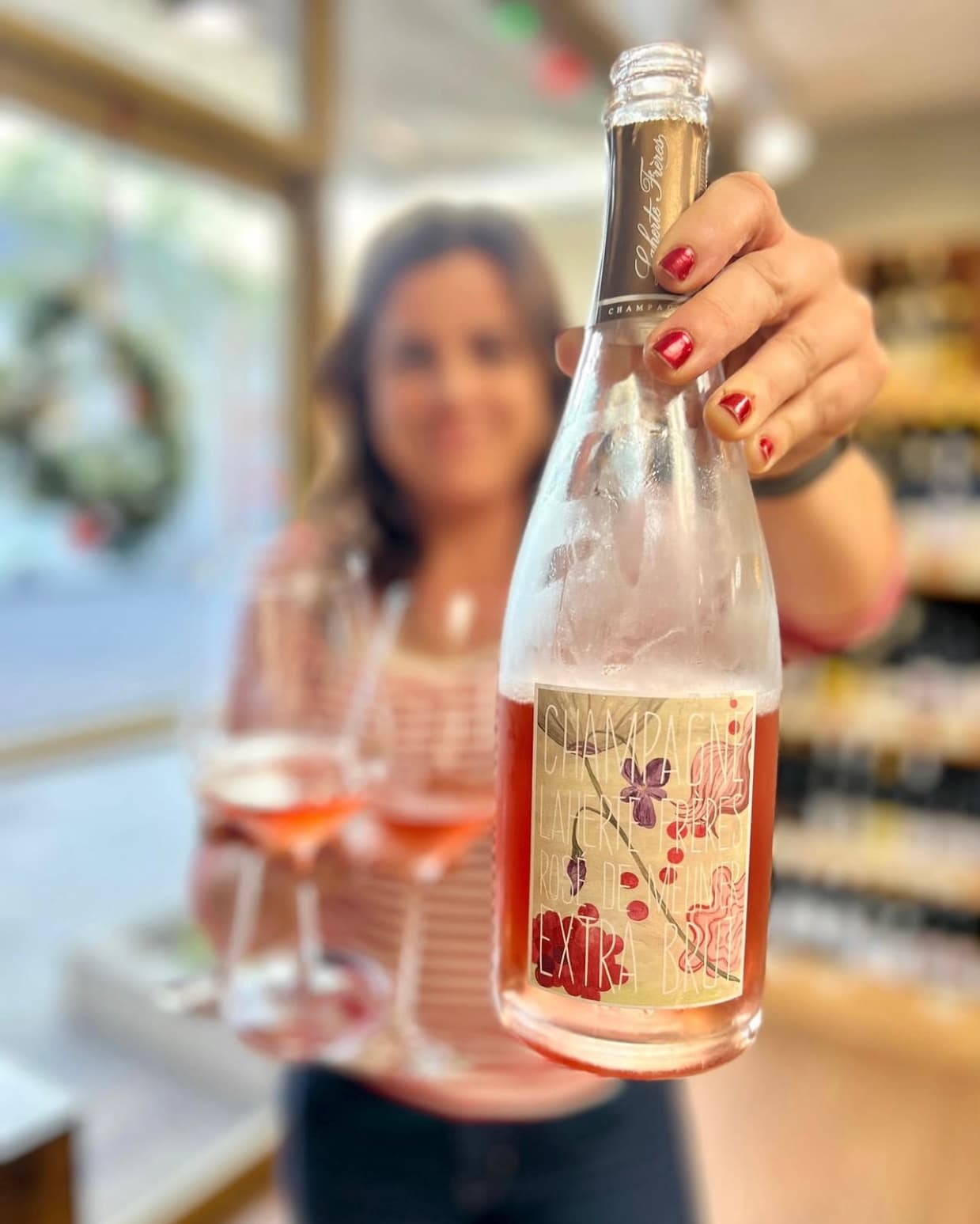 A person holding a chilled bottle of champagne in the foreground, with a blurred background showing wine glasses and a shop interior.
