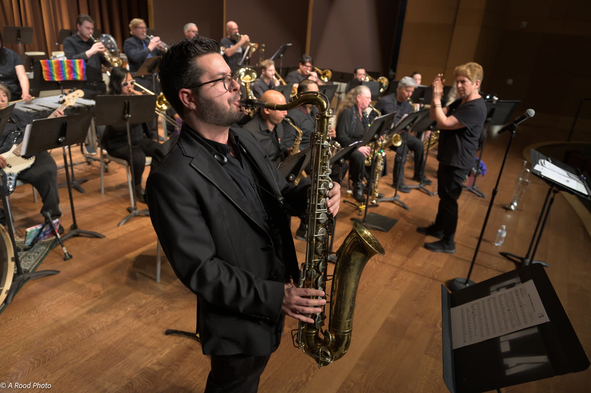 A sax player stands on stage in the foreground while a conductor leads a band in the background.