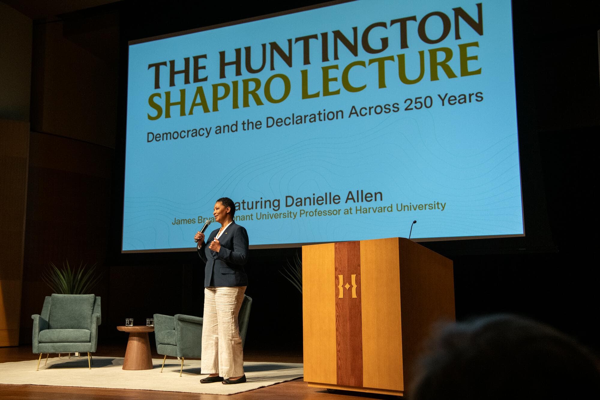 A person in a dark jacket and cream slacks speaks into a microphone on a stage in front of a large screen that says "The Huntington Shapiro Lecture, Democracy and the Declaration Across 250 Years, Featuring Danielle Allen.":