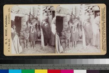 A Hopi woman holds a child in front of a ladder at an unidentified pueblo