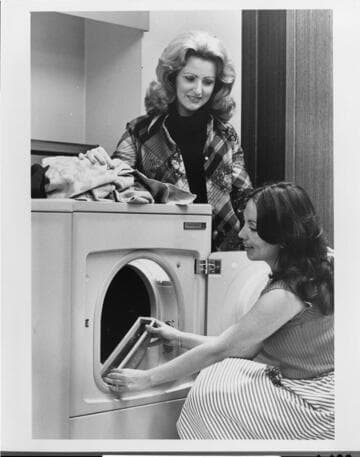 Two women stand at the family drier as one cleans out the drier lint trap to conserve energy
