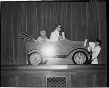 Performance of skit at Edison auditorium. Man and woman in cardboard car with another man fixing a flat on the car
