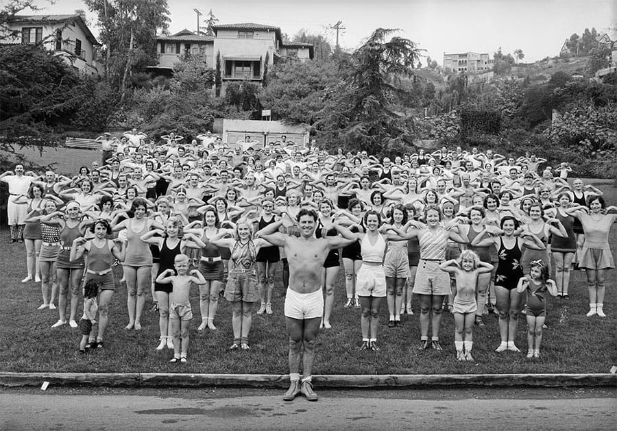 A group of 100 or so people in bathing suits and shorts, make a "crab" pose, showing off the muscles in their arms.