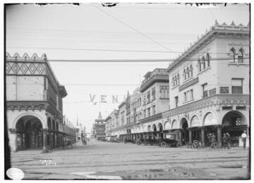 An area view of Windward Avenue in Venice, California, [showing a Turkish Bath House, theaters, an apartment house and automobiles common to 1914 parked along the street. There is a large sign at the intersection that reads: VENICE. People are socializing along the street and there is a horse