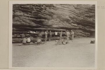 Camp on right bank at Mile 115. Seymour and Jordan seated left, Joe Desloge kneeling, MacDonald and Marston center, Sanderson in back of groceries, Taylor and Desloge at extreme right