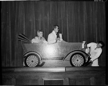 Performance of skit at Edison auditorium. Man and woman in cardboard car with another man fixing a flat on the car