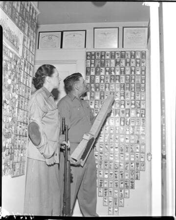 Man and woman in room filled with framed awards and medals for marksmanship. Woman is holding a sharpshooters rifle and man is holding a blank un