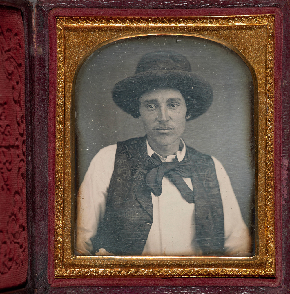 A golden-brown frame with red velvet accents surrounds a grayscale portrait of a man in a white shirt, with a black tie, vest, and hat.