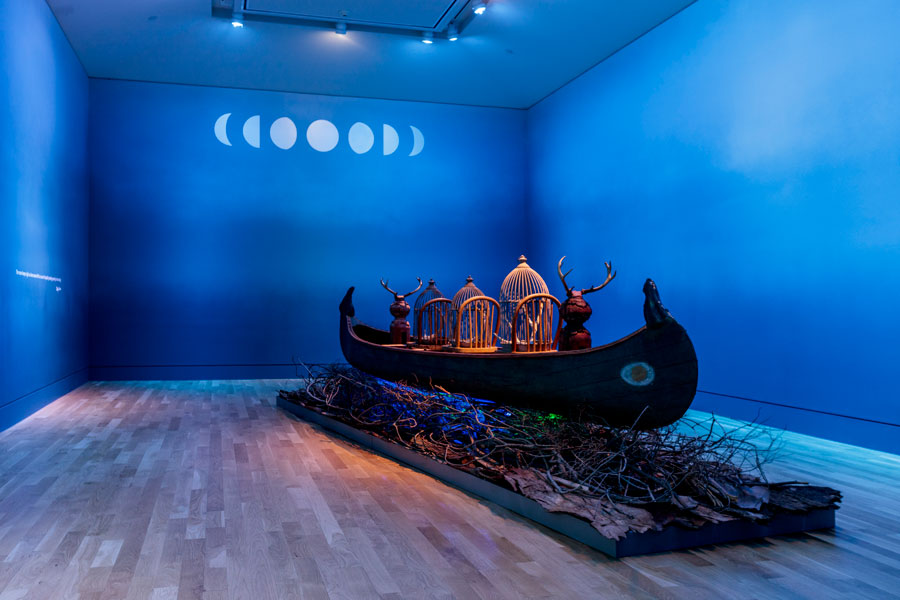 A wood canoe sits on top of a bed of dry branches in a blue room with phases of the moon painted on the wall.