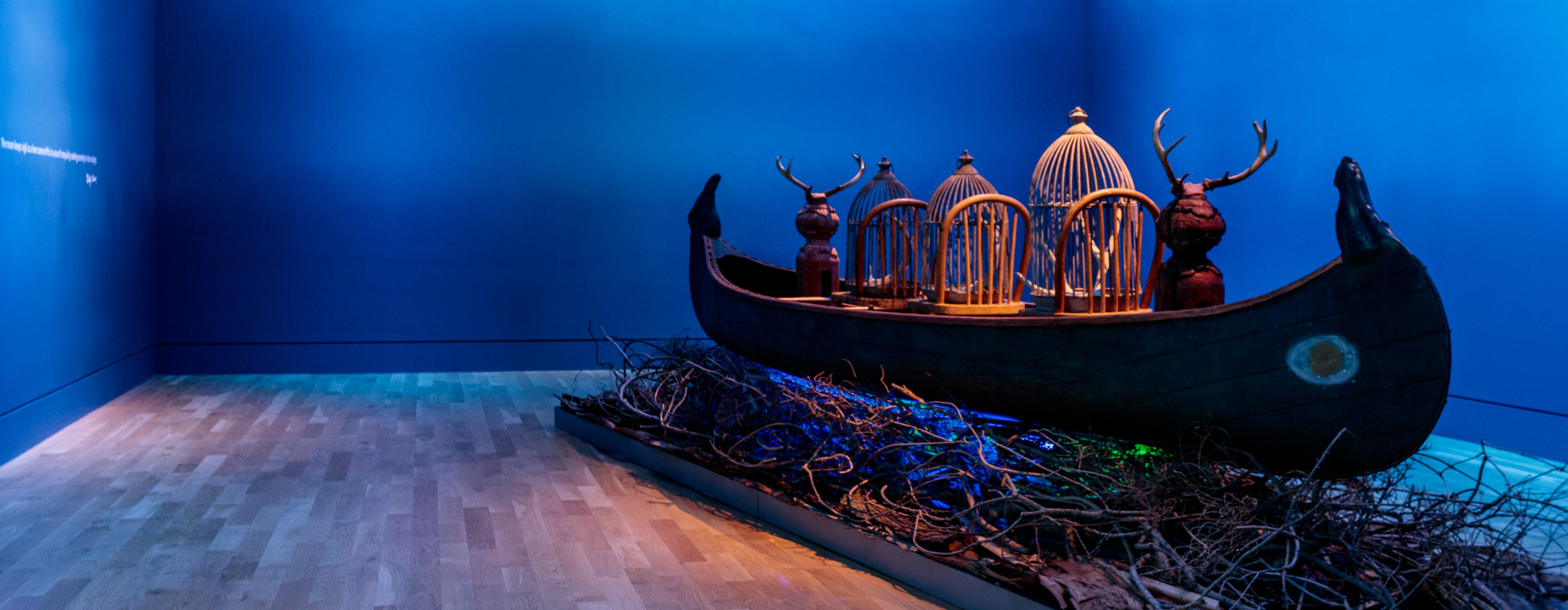 A canoe with wooden objects inside floats over a bed of dry branches in a blue room.