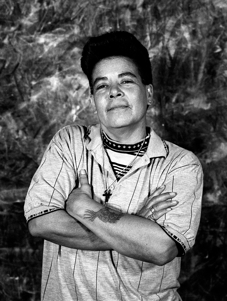 Laura Aguilar, #13, from the series Plush Pony, 1992 Queer middle-aged woman poses with her arms crossed in front of a roughly textured backdrop