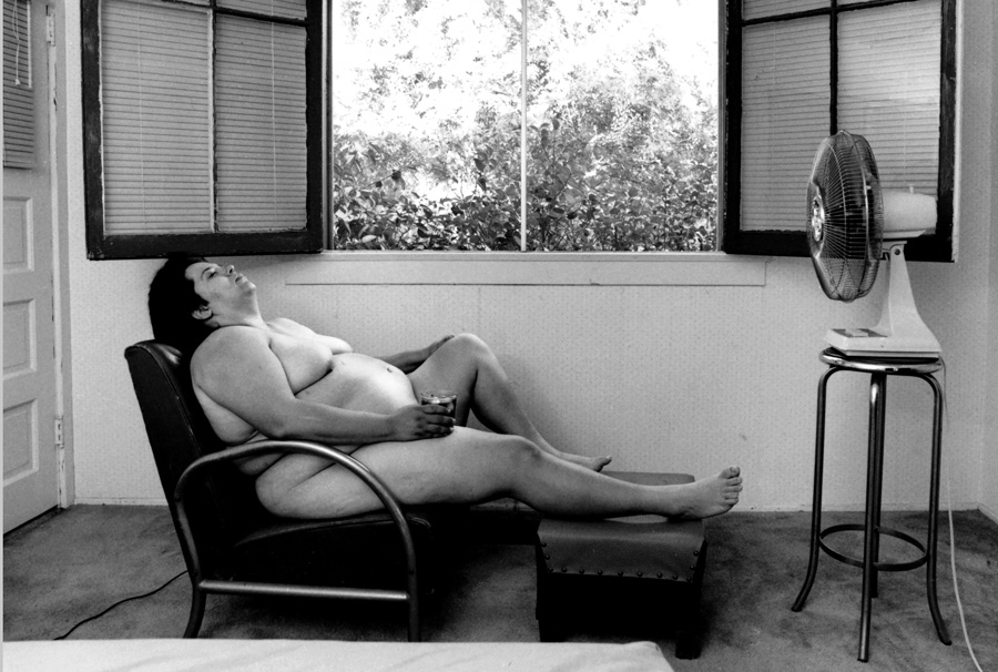 Laura Aguilar, In Sandy’s Room, 1989, printed 1990 Obese woman sitting naked in front of fan and open windows with a drink in her hand